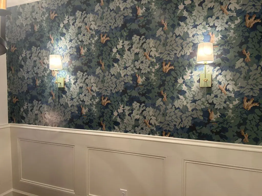 Dining room with dark blue-green botanical wallpaper above white wainscot panels, lit by two brass sconces