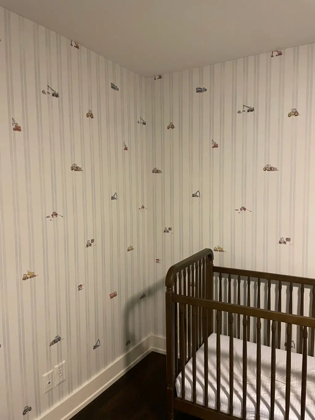 Nursery corner papered in a pale ticking stripe with small scattered vintage-car motifs, beside a dark wood crib